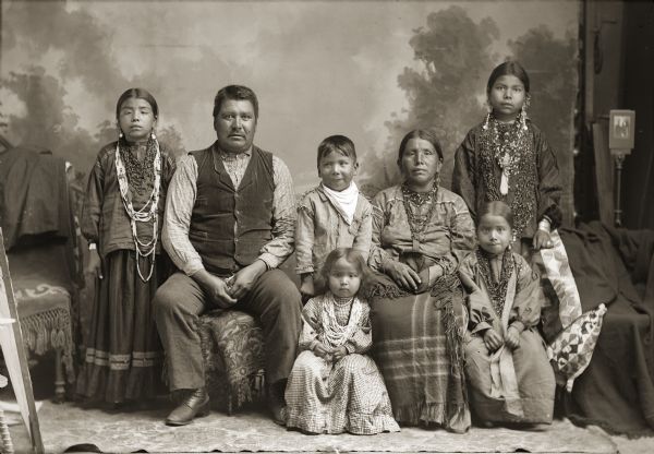 Full-length studio portrait of a Ho-Chunk family, including a man, woman, four girls, and a boy posing in front of a painted backdrop. The woman and older girls are wearing degrees of regalia, and the remaining individuals are wearing contemporary dress. The Whitebear family was referred to in the census rolls with the last name of White but they used the name Whitebear. From left to right are Ella Whitebear (MauNaPayWinKah), father Jim Whitebear (HoonchSkaKah), Dan Whitebear (HeNukKah), mother Kate Whitebear (MawChePauSayWinKah), and Daisy Whitebear (WeHunKah). Lucy Whitebear (HoDaHooKah) sits in front of Dan, and Viola Whitebear (UkSeUkKah) is sitting in front of Daisy.