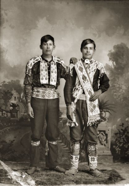 Full-length studio portrait of Frank Winneshiek (WaConChaHoNoKah), left, and Henry Winneshiek (WaConChaRooNayKah), right, in front of a painted backdrop. Both men are wearing traditional beaded shirts, garters, and Ho-Chunk-style moccasins. Frank is wearing two bandolier bags and a bone choker with a shell. Traditionally this choker was worn as armor to protect the throat during battle.