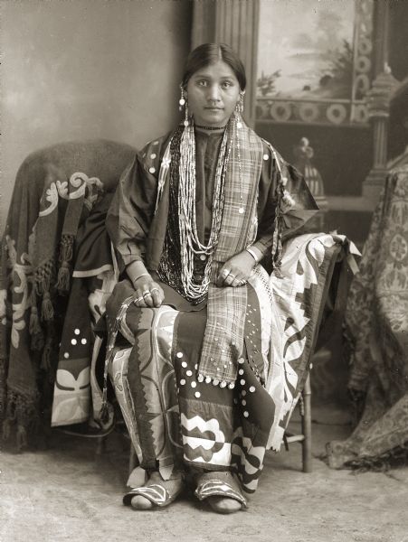 Studio portrait in front of a painted backdrop of Flora Thundercloud Funmaker Bearheart (WaNekChaWinKah), daughter of Moheek Thundercloud (MaZheeWeeKah) and Annie Blowsnake Thundercloud (WaConChaSkaWinKah). Flora is sitting in a chair covered by a silk appliqué blanket (wai) and is dressed in a fine appliqué dress (wawaje) with silver dimes attached to the skirt. The plaid ribbon with silver coins hanging on her left side, and the strip of side-stitch beadwork (paque) hanging over her right shoulder would have been worn down the back extending from her hair wrap, but they have been brought to the front for the photograph. Small silver coins accent her blouse. The flaps of Flora’s moccasins (waukoocharah) are decorated in silk appliqué to match her dress. Many Ho-Chunk moccasins of this style have the top flaps decorated with beadwork.