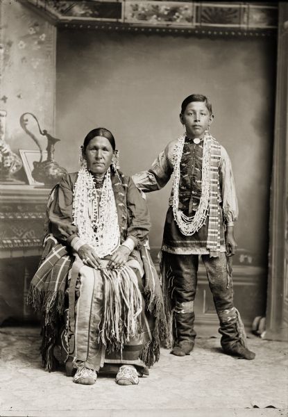 Full-length studio portrait of a Ho-Chunk woman posing sitting and a Ho-Chunk boy posing standing in front of a painted backdrop. Susie Kingswan (HeNuKah) is wearing a fine pair of appliqué beaded Ho-Chunk moccasins. Her son Fred Kingswan (MaHeNoGinKah) is wearing traditional leather leggings (waguca) and beaded garters.
