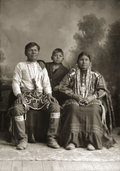 Full-length studio portrait of a Ho-Chunk man, woman and child posing sitting and standing in front of a painted backdrop. They are identified as Moheek Thundercloud (MaZheeWeeKah) and Annie Blowsnake Thundercloud (WaConChaSkaWinKah), sitting, with their son, Benjamin Raymond Thundercloud (NySaGaShiskKah) standing behind them in the center.