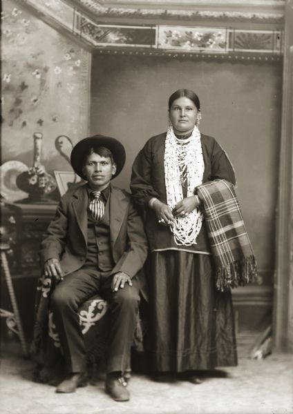 Full-length studio portrait in front of a painted backdrop of a Ho-Chunk man posing sitting and wearing a suit jacket, tie, and hat. Next to him on the right is a Ho-Chunk woman posing standing and holding a wool shawl, and wearing bead necklaces and long earrings. Henry Thunder (WahQuaHoPinKah) and Soldier Woman (MahNaPayWinKah).