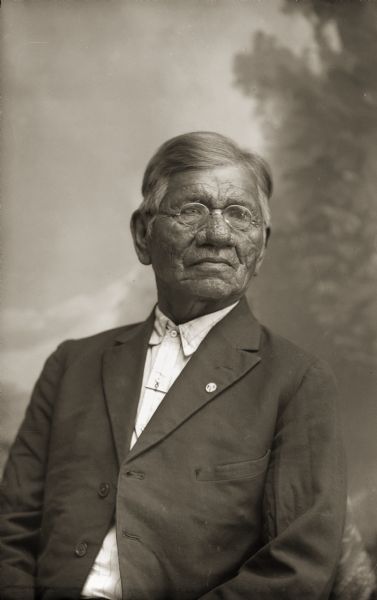 Waist-up studio portrait of an elderly Ho-Chunk man posing sitting in front of a painted backdrop. He is wearing a suit jacket and eyeglasses, and is identified as Moheek Thundercloud (MaZheeWeeKah) as an older man. It was very rare for Ho-Chunk to wear eyeglasses in Van Schaick’s photographs.