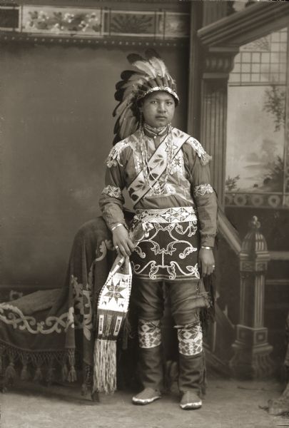 Full-length studio portrait of a young Ho-Chunk man posing in front of a painted backdrop. Benjamin Raymond Thundercloud (NySaGaShiskKah), son of Annie Blowsnake Thundercloud (WaConChaSkaWinKah) and Moheek Thundercloud (MaZheeWeeKah), is wearing a mix of Ho-Chunk and regalia from other tribes. The eagle feather bonnet, commonly referred to as a war bonnet (maasuu wookanak), was not traditionally worn by the Ho-Chunk but was adopted from the Plains tribes after the Ho-Chunk participated in Wild West shows. His shirt (woonazi) is a traditional Ho-Chunk beaded shirt with loom-beaded strips sewn on the shoulders and the upper front of the shirt. The beaded armbands (ahparrah), beaded belt (pecoonrah), garters (hoowayescurrah), and breechcloth (tauneeodoorah) are Ho-Chunk. The pipe bag and moccasins are a Plains Indian style.