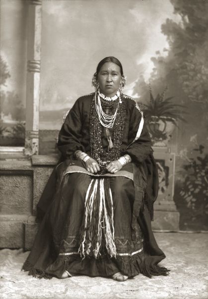 Full-length studio portrait in front of a painted backdrop of a Ho-Chunk woman posing sitting on a prop stone wall. She is wearing full regalia, including many necklaces, earrings, and fringed dress. She is identified as Lucy Long-Wolf Winneshiek (ShunkChunkAWinKah).
