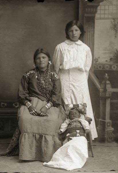 Full-length studio portrait in front of a painted backdrop of two young Ho-Chunk women and a Ho-Chunk infant. The woman sitting on the left is wearing regalia, and the woman standing on the right is wearing contemporary clothing. The infant is in a papoose on the floor between the two women. They are identified as Sarah B. Windblow Thundercloud (HeChoWinKah), sitting next to her sister Nellie Windblow Decorah (HaWePinWinKah), who is dressed in a white school uniform, ca. 1909. Sarah’s son Lawrence Leonard Thundercloud (CooNooKah), is sleeping comfortably in a Ho-Chunk cradleboard.