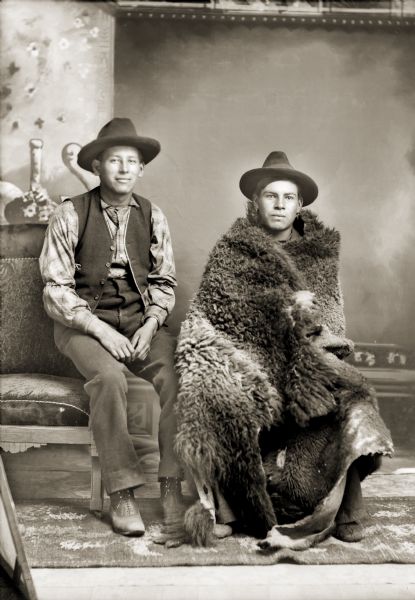 Full-length studio portrait in front of a painted backdrop of two Ho-Chunk men posing sitting. They are wearing hats, and the man on the left, Henry Winneshiek, is wearing a vest, plaid shirt, and trousers. The man on the right, Frank Winneshiek, is wrapped in a buffalo skin/robe and is wearing earrings.
