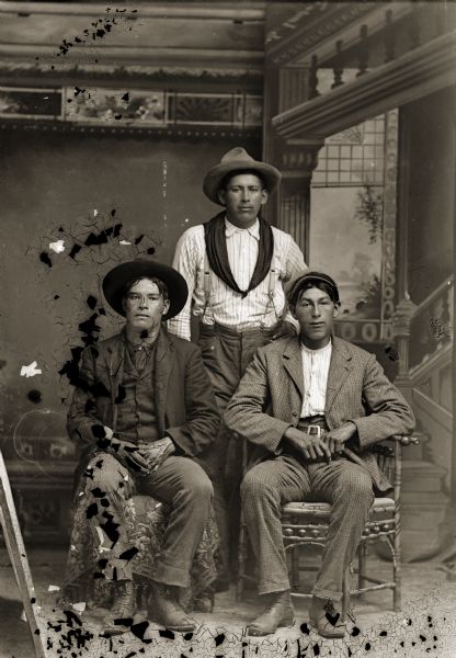 Full-length studio portrait in front of a painted backdrop of two Ho-Chunk men posing sitting in front of a Ho-Chunk man posing standing The men are all wearing hats, and the two men sitting are wearing suits. Identified from left to right as, Henry Winneshiek (WaConChaRooNayKah), Frank Winneshiek (WaConChaHoNoKah), and Richard (Luther) Winneshiek (MaHeKeeZheeNaKah).