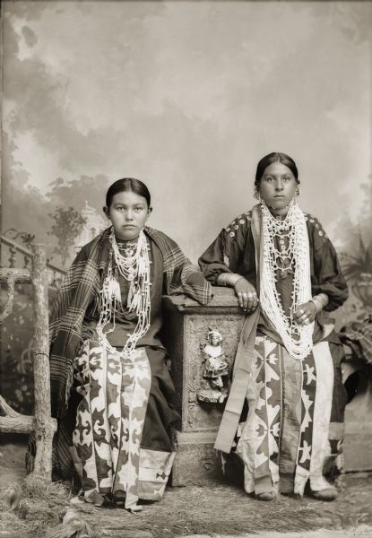 Studio portrait in front of a painted backdrop of two young Ho-Chunk women posing sitting on either side of a prop stone wall and prop wood fence. They are wearing several necklaces with ribbon work skirts. The woman on the left is wearing a plaid shawl over her shoulders, and the woman on the right is wearing several rings, earrings, and file bracelets. They are identified as Emma Bigbear (WaConChaZeeWinKah) left and Lillie Winneshiek (WaConChaWinKah).