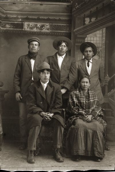 Studio portrait of group of people posing in front of a painted backdrop. A Ho-Chunk man on the left is wearing a suit and overcoat, and a Ho-Chunk woman on the right is wearing a shawl and dark-colored dress. Three Ho-Chunk men are standing behind them wearing suits and hats. They are identified as John Blackhawk (NoJumpKah), seated to the left of Lucy Long-Wolf Winneshiek (ShunkChunkAWinKah). Standing left to right are Henry Winneshiek (WaConChaRooNayKah), Edward Winneshiek (NaZhooMonEKah), and George Lonetree (HoonchXeDaGah).