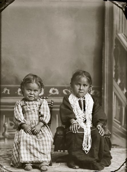 Studio portrait in front of a painted backdrop of two small Ho-Chunk girls posing sitting in chairs. The girl on the left is wearing a few necklaces, moccasins, and a plaid dress, and the girl on the right is wearing many necklaces, earrings, file bracelets, and moccasins. They are identified as Maude Lookingglass Browneagle (ENooKah) on the right and her sister Liola Browneagle (WeHunKah), the daughters of Frank Browneagle (HeWaKaKayReKah) and Annie Snake (KhaWinKeeSinchHayWinKah). Maude reportedly died in 1970 and her sister sometime afterward.