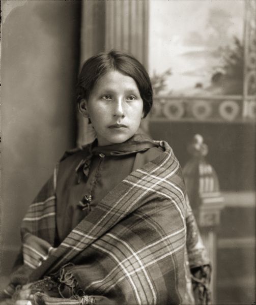 Waist-up studio portrait of a young Ho-Chunk woman posing sitting. She is wrapped in a plaid fringed shawl, and is identified as Kate Thunder Miner (WaRoSheSepEWinKah).