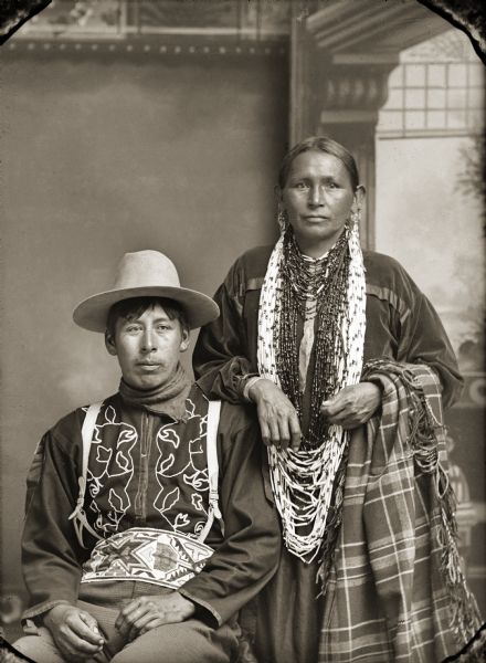 Studio portrait in front of a painted backdrop of a Ho-Chunk man and woman. Edward Winneshiek (NaZhooMonEKah), is wearing a rare floral hand-beaded Ho-Chunk shirt and loom-beaded belt, and is sitting next to Annie Lucy YankeeBill (HaTaHeMonEWinKah).
