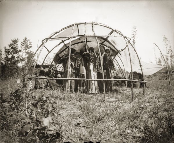 A group of Ho-Chunk people standing in the rear of a Buffalo Dance Lodge. The Buffalo Dance would take place in the spring, when the grass was long enough to tie in a knot.