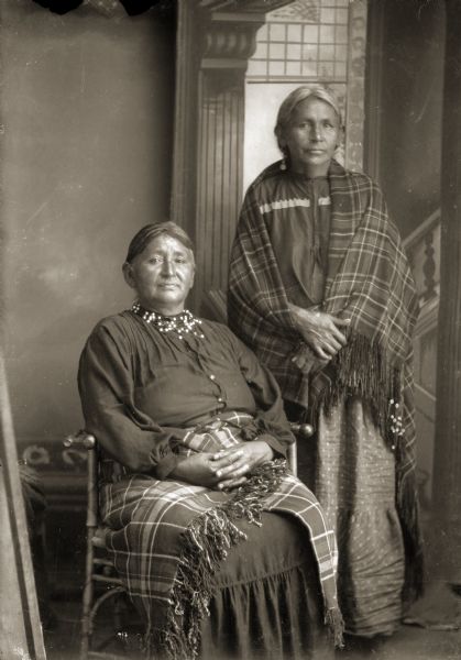 Studio portrait of two older Ho-Chunk women posing in front of a painted backdrop. The woman on the left, Rachel Funmaker Winneshiek (HockJawKooWinKah), is sitting and is wearing a shawl around her waist, and a necklace. The woman standing on the right, wearing a shawl around her shoulders, is identified as Mary Thunderchief Snowball (WaSayMaKaWinKah).