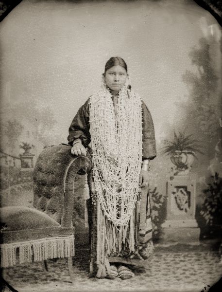 Full-length studio portrait of Annie Blowsnake Thundercloud (WaConChaSkaWinKah), posing standing in front of a painted backdrop. She is wearing many necklaces, and other jewelry including earrings, file bracelets, and rings. Her moccasins have ribbon trimmed flaps and her right arm is resting on a velvet fringed chair. Annie had just completed a ribbon work (silk appliqué) dress (wawaje) when she stopped in at Van Schaick’s studio. Moments before the photograph was taken, Annie’s mother draped several large necklaces of white bugles and shells around her neck, completely covering her appliqué. Annie’s face registers her disappointment. Fine regalia complements the ensemble: silver bracelets (aipa), earrings (nachahocpok), rings (naap hirusgic), and traditional Ho-Chunk moccasins (waukoocharah). Photographs of this type were made into postcards and sent to relatives in other Ho-Chunk settlements to serve as notice to eligible young men of the young woman’s appearance and dowry in wampum (worookshigerah) and jewelry.