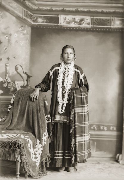 Full-length studio portrait in front of a painted backdrop of a Ho-Chunk woman posing standing with her right arm on a chair draped with a cloth. She is wearing a Winnebago charm, coin earrings, a bird bone, a wampum necklace, and holding Van Schaick's studio blanket over her left arm. Identified as Emma Blackhawk Bigbear Beaver-Smith Holt (Female Yellowthunder) (WauKonChawZeeWinKah), married to Henry Holt.