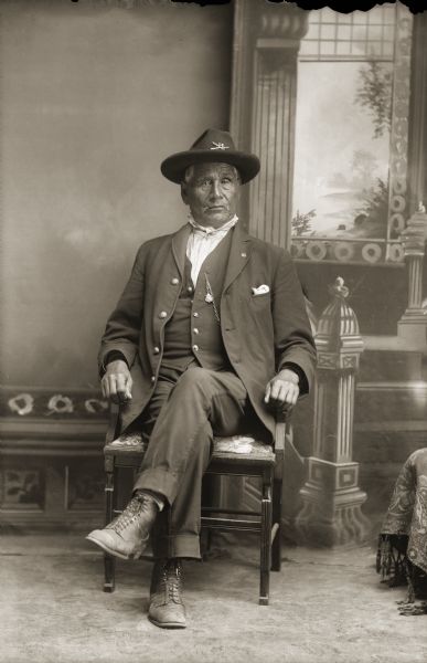Full-length studio portrait of an elderly Ho-Chunk man posing sitting. He is wearing a suit jacket, vest with watch fob, trousers, and a hat with a United States army insignia. Identified as Civil War veteran James Bird (WeeJukeKah or MaKeSucheKaw). The emblem on his hat is made of two crossed arrows, the insignia of a Civil War Indian scout. James Bird was a private with Company A of the Omaha Scouts, a Civil War outfit consisting of 6 white officers and 72 Ho-Chunk soldiers.