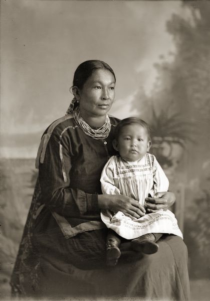 Studio portrait of a Ho-Chunk woman posing sitting and holding a small child in her lap front of a painted backdrop. Martha Lyons-Lowe Stacy (KaRaChoWinKah) is holding her son James Stacy (NaHeKah). James, son of John Stacy (ChoNoKayHunKah), died at the age of two.