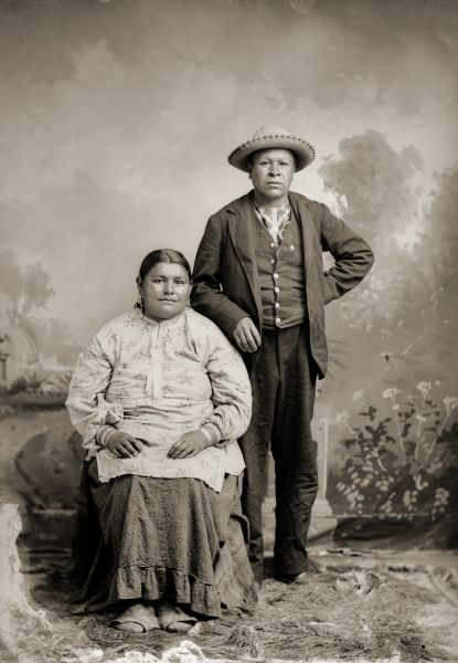Full-length studio portrait of a Ho-Chunk man posing standing and a Ho-Chunk woman posing sitting in front of a painted backdrop. They are identified as Liddy Walkingcloud Winneshiek (KsoonchEMonEWinKah), and Little (Nady) Winneshiek (NoGinKah) in contemporary clothing.