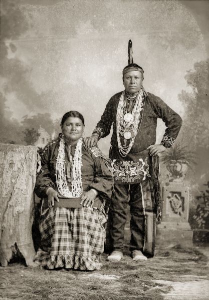Full-length studio portrait of a Ho-Chunk man posing standing and a Ho-Chunk woman posing sitting in front of a painted backdrop and near a tree stump. They are identified as Liddy Walkingcloud Winneshiek (KsoonchEMonEWinKah) and her husband, Little (Nady) Winneshiek (NoGinKah) in traditional clothing. Little Winneshiek is wearing a finger woven sash and eagle feather on his head, and an "unofficial" George Washington Indian peace medal with a crude likeness of Washington around his neck.