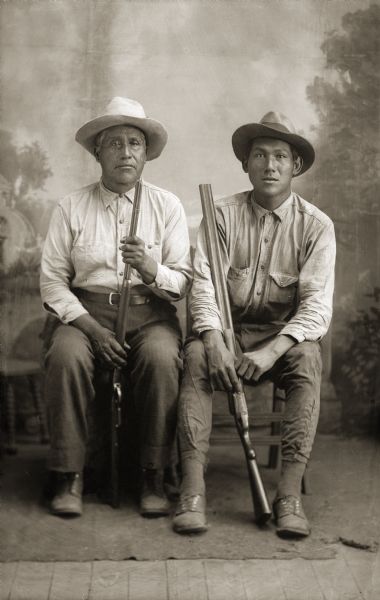 Full-length tudio portrait of Ho-Chunk men, Charles Sine Sr. and Harry Swallow, both posing sitting, wearing hats, and holding guns in front of a painted backdrop. The older man on the left, Charles (Charlie) Sine Sr., is holding a repeating rifle. The younger man on right, Harry (Henry) Swallow from Nebraska, is holding a shotgun.