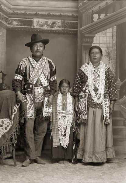 Full-length studio portrait in front of a painted backdrop of a Ho-Chunk man, woman, and girl, all posing standing. They are identified as David Goodvillage (WauHeTonChoEKah) on the left, Lucy Blackhawk Bighead Davis Goodvillage (NeAhTakEWinKah) on the right, and Lucy’s granddaughter Mabel Blackhawk St. Cyr, in the center. During this period it was typical for the women to wear all traditional clothing, and the men to wear a mix of contemporary and traditional.