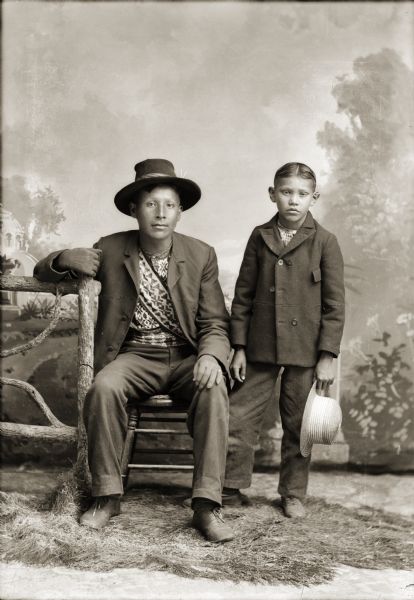 Studio portrait in front of a painted backdrop of a young Ho-Chunk man posing sitting in a chair near a prop fence, wearing a suit jacket, hat, and bandoleer, and a Ho-Chunk boy posing standing wearing a pea coat and holding a hat in his left hand. Will Stohega Carriman (WonkShiekStoHeGah), sitting next to Andrew John (Big) Blackhawk (WaConChaHoNoKah), wearing a beaded belt and bandoliers across his chest.