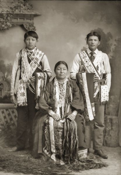 Mary Goodvillage Hindsley (ChoWasSkaWinKah) is sitting in front of Henry Hindsley (MaHePeWinKah), left, and Henry Badsoldier Stacy (MonNawPaeSheSheckKaw). Mary is dressed in traditional Ho-Chunk clothing, including a silk appliqué skirt (wawaje), necklaces (wanapi), German-silver bracelets (zuura sgaa aipa), and a finger-woven wool sash. Both young men are wearing beaded Ho-Chunk bandolier bags and bone and shell bandoliers and are holding silk appliqué blankets. These bandolier bags are of Ho-Chunk design. The Ho-Chunk did trade for bandolier bags with the Ojibwa, a common practice at this time. It is said that the price for a bandolier bag was one good horse.