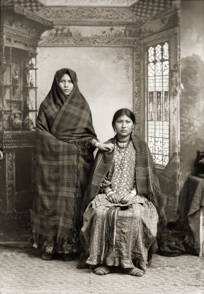 Studio portrait in front of a painted backdrop of a Ho-Chunk woman posing standing on the left wearing rings and file bracelets, and wrapped in a plaid shawl, only showing her face and her left arm which is resting on the other woman's shoulder. Sitting on the right is a Ho-Chunk woman, Red Bird Flying, the wife of Frank Lincoln, wearing a dress (hinukwaje) made of fabric with an elaborate print, several necklaces, earrings, file bracelets, and rings. Women's dresses were typically made of solid or simple print fabric.