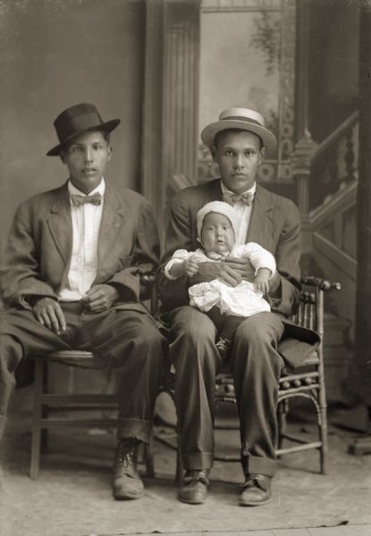 Studio portrait in front of a painted backdrop of two Ho-Chunk men posing sitting and wearing suits, bow ties, and hats. Andrew John (Big) Blackhawk (WaConChaHoNoKah) holding his son Clarence Blackhawk (WaCheeWaHaKah) and sitting next to an unidentified man.