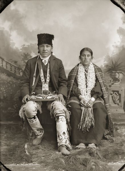 Studio portrait of a Ho-Chunk man and woman posing sitting in front of a painted backdrop. They are identified as Ed Greengrass (CheWinCheKayRayHeKah) and his wife Jennie (Johnnie) Blackhawk (CheNunkMonEWinKah) in their finest Ho-Chunk clothing. Ed is wearing an otter-fur turban and floral appliqué beaded aprons and leggings.