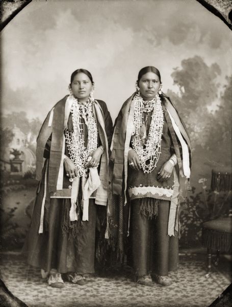 Full-length studio portrait in front of a painted backdrop of two unidentified women in traditional clothing with traditional necklaces and strips of silk ribbon over their shoulders. Both have ribbon work on the bottom edge of their blouses. The woman on the left has a slit on the front of her blouse with ribbons sewn on both sides. This is a style used by some of the women from Nebraska.