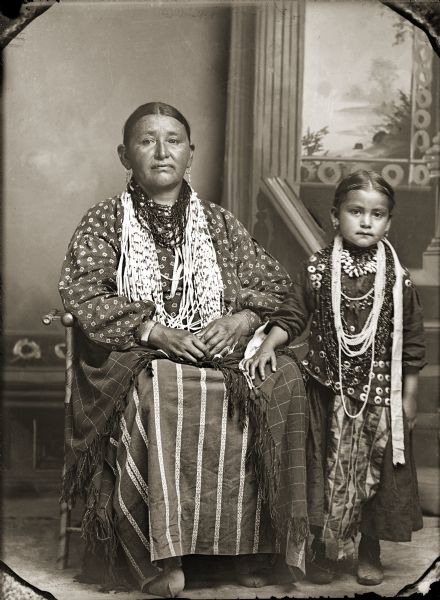 Studio portrait of a Ho-Chunk woman and girl wearing regalia posing in front of a painted backdrop. The woman sitting on the left is wearing several necklaces, earrings, file bracelets, and a shawl wrapped around her waist. The Ho-Chunk girl is standing on the right with her right hand on the woman's left knee. She is wearing several necklaces, a traditional blouse, earrings, streamers, ribbon-work skirt, and moccasins. They are identified as Rachel Funmaker Winneshiek (HockJawKooWinKah), left, sitting next to Fannie Winneshiek (HoChunkEHoNoNeekKah) (Queen Redwing).