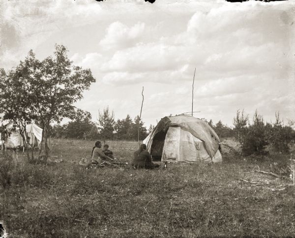 Three unidentified Ho-Chunk people and a dog are sitting outside of their canvas-covered ciiporoke. A summer house can be seen through the trees.