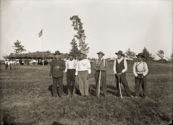Six Ho-Chunk men lined up for a group portrait at the powwow grounds. The men with sticks in their hands are members of the Bear Clan, who act as police for the tribe and handle security at tribal events. From left to right are Andrew Bigsoldier (WaConChaShootchKah), Henry Winneshiek (WaConChaRooNayKah), George Monegar (EwaOnaGinKah), Albert Thunder (CheNunkEToKaRaKah), Frank Washington Lincoln (ChakShepKonNeKah), and Max Bearheart (MoNeKah).