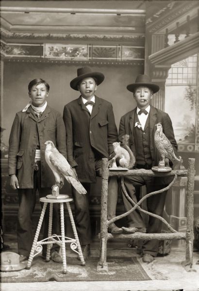 Full-length studio portrait of three young Ho-Chunk men wearing suit jackets, trousers, and neckties in front of a painted backdrop. Two are wearing hats and smoking, and have their feet resting on a prop fence. Identified from left to right as, Henry Greencrow (CooNooZeeKah), William Hall (HunkKah) and Charlie Greengrass (HoeHumpCheeKayRayHeKah), posing with two stuffed birds and a stuffed squirrel.