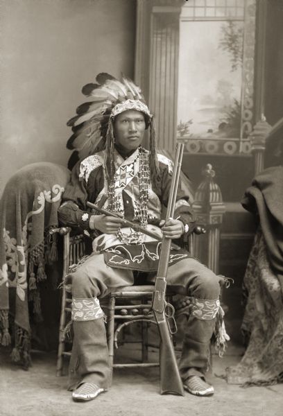 Full-length studio portrait of a World War I Ho-Chunk veteran, James George Hanakah (PaitchDoAhHeKah), wearing full Ho-Chunk regalia, except for his Plains-style moccasins. The eagle feather bonnet was not traditionally Ho-Chunk but was adopted by the late 1800s after they participated in the Wild West shows. He is sitting on a chair in front of a painted backdrop, holding a rifle and tomahawk.