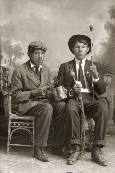 Full-length studio portrait of two Ho-Chunk men posing sitting in chairs in front of a painted backdrop. Sam Thunderking Lowe (HoChumpHoNoNikKah), on the left, and Julius Whitedog (ChakShepWasKaHeKah), on the right. Both men are holding items used in the Native American Church. Sam holds a can used as a drum, and Julius has a staff and gourd rattle.