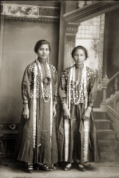 Full-length studio portrait in front of a painted backdrop of two young Ho-Chunk women posing standing. They are identified as Flora Thundercloud Funmaker Bearheart (WaNekChaWinKah), left, and Mary May Thundercloud Deer (WaNeekPiWinKah). They are both wearing blouses covered with small silver brooches (heapoke), and contemporary shoes. Blouses covered with heapoke were sometimes referred to as “buckle blouses” because the brooches looked like miniature buckles.