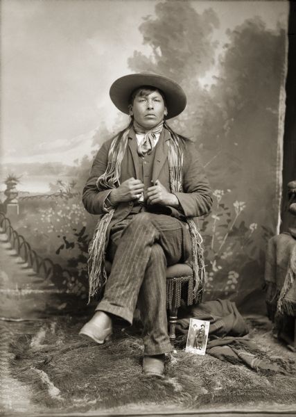 Studio portrait in front of a painted backdrop of an unidentified Ho-Chunk man with long hair posing sitting with his legs crossed. He is wearing a suit, bandana, finger woven sash over his shoulders, and a hat. Propped up by a shawl on the floor is a cabinet card from Van Schaick's studio with a picture of two Ho-Chunk men.