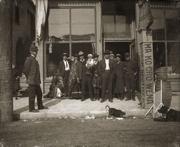 A group of Ho-Chunk gathering in front of Werner Drugstore on Main Street. The signpost on the right side indicates that English was not the first language of many of the area’s residents. One side of the post gives the Norwegian for drug store, “APOTHEK.” The front side reads “MA KO CRO WENA,” which means “medicine sold here” in Ho-Chunk. Thomas Thunder (HoonchHaGaKah) is standing on the left and Hester Decorah Lowery (NoGinWinKah) is sitting next to the building.