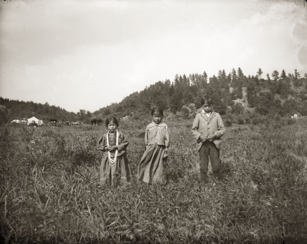 Three Ho-Chunk children posing for a group portrait in a field known as Browneagle Bottoms. The children are, left to right, Maude Browneagle (WeHunKah), Liola Browneagle (ENooKah), and Jesse Stacy (MoRoJaeHooKah). The boy standing on the right is wearing a suit coat, while the two girls in the center and left and are wearing dresses. The girl posing on the left is wearing several necklaces. In the background are horses, wagons, and a tent, and low hills with trees.
