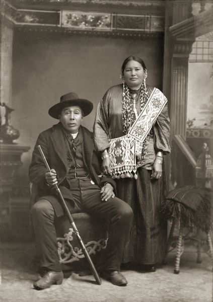 Full-length studio portrait of a Ho-Chunk man, George Greengrass and his wife in front of a painted backdrop. George Greengrass (WauKeCooPeRayHeKah) and Emma Lookingglass Greengrass (ChayHeHooNooKah). Emma is standing and wearing a bandolier with an offset top strap which was common to Ho-Chunk bandolier bags. He is the grandfather of LaVern Carriman. He is sitting holding a rifle and is wearing a hat, suit jacket, vest with watch fob, scarf, and trousers.