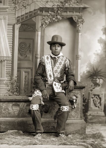 Full-length studio portrait of David Bow Littlesoldier (MauJchayMauNeeKah) posing sitting on a prop stone wall in front of a painted backdrop that appears in many of Van Schaick’s photographs. He is wearing a hat (wookanak), a beaded shirt (woonazi), two bandolier bags crossed over his chest, loom-beaded garters, and traditional Ho-Chunk moccasins (caahawaguje).