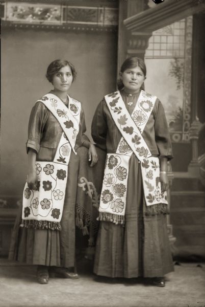 Full-length studio portrait of Flora Thundercloud Funmaker Bearheart (WaNekChaWinKah), left, and Sarah B. Windblow Thundercloud (HeChoWinKah), right. Both women are wearing floral beaded bandolier bags across their shoulders.