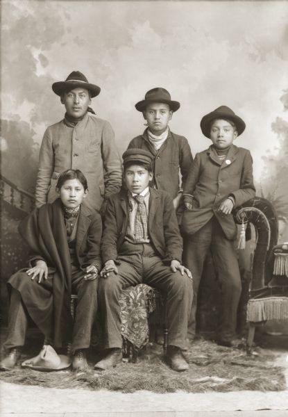 Full-length studio group portrait of young Ho-Chunk men. Standing (l to r) are George Otter (Hay Cho Kah), Johnnie Thunder (Hoonk Nee Kah), and George Bearchief. Sitting (l to r) John Rainbow and an unidentified young man (name illegible), and are holding cigars. All the men are wearing hats except the boy sitting on the left who has a hat at his feet. In the background is a painted backdrop.