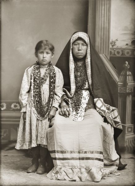 Studio portrait of Ho-Chunk sisters, Nellie Winneshiek Twocrow Redcloud, (WaConChaWinKah) and her older sister, Kate Winneshiek Lonetree (WauKonChawKooWinKah). Nellie is posing standing on the left, and is wearing several necklaces and a light-colored print dress. Kate is posing sitting on the right, and is wearing several necklaces, rings, file bracelets, and a dark-colored ribbon work shawl over her head.
