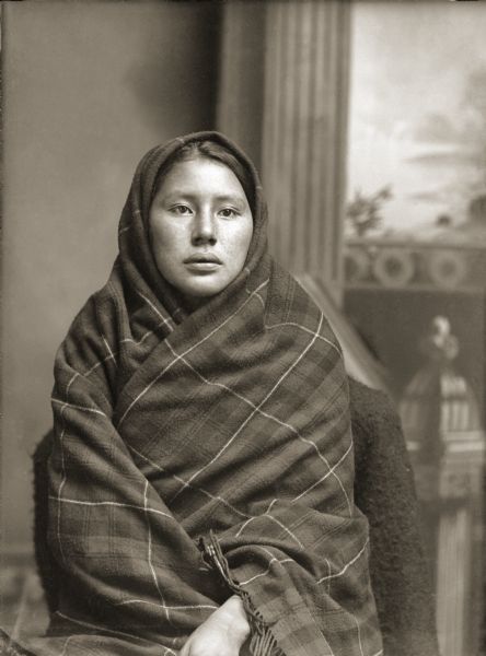 Waist-up studio portrait of a young Ho-Chunk woman posing sitting in front of a painted backdrop, wrapped in a plaid fringed shawl showing only her face, a style displayed in a number of Van Schaick's photographs of this period. She is identified as Grace Decorra Clay Whitegull (BeekSkaWinKah).