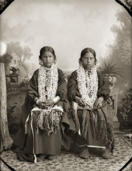 Full-length studio portrait in front of a painted backdrop of two women posing sitting. Emma Mary White Greencloud-Redcloud Thunder (JumpBroShunNupChunTinWinKah), left, and Sally Dora Redbird Goodvillage Whitewater (WauNeekShootchWinKah).